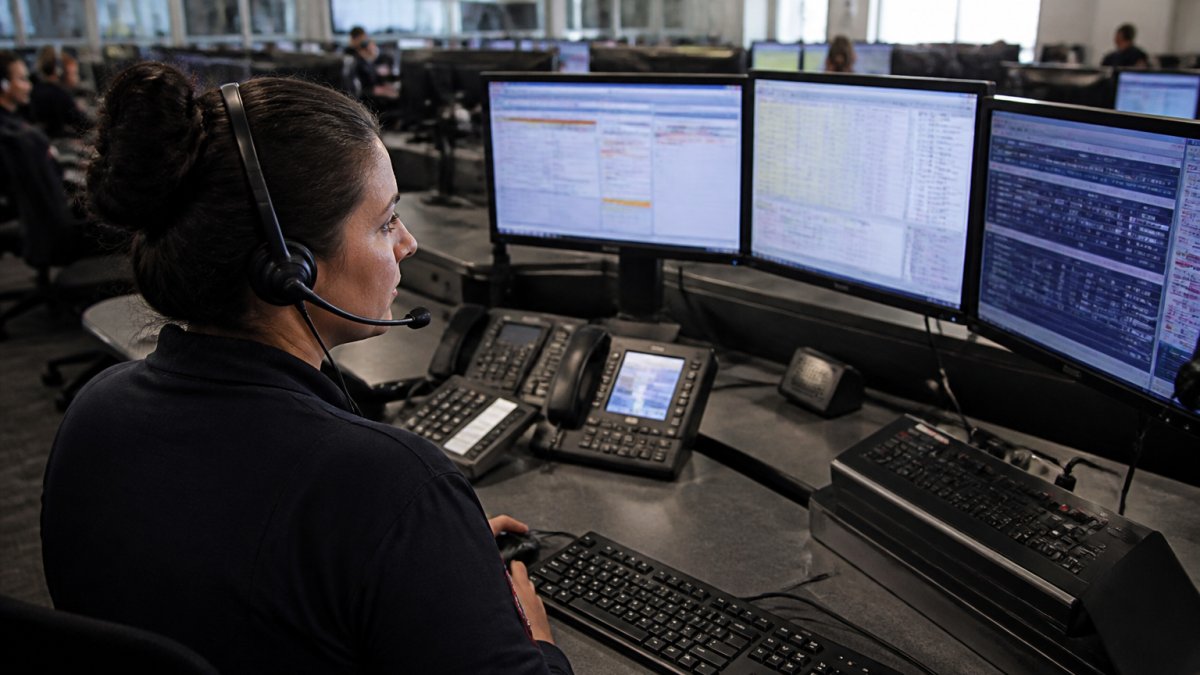 Professional emergency dispatcher at workstation monitoring multiple screens displaying call data, CAD system, and AI-assisted incident information in modern dispatch center with collaborative workflow visible