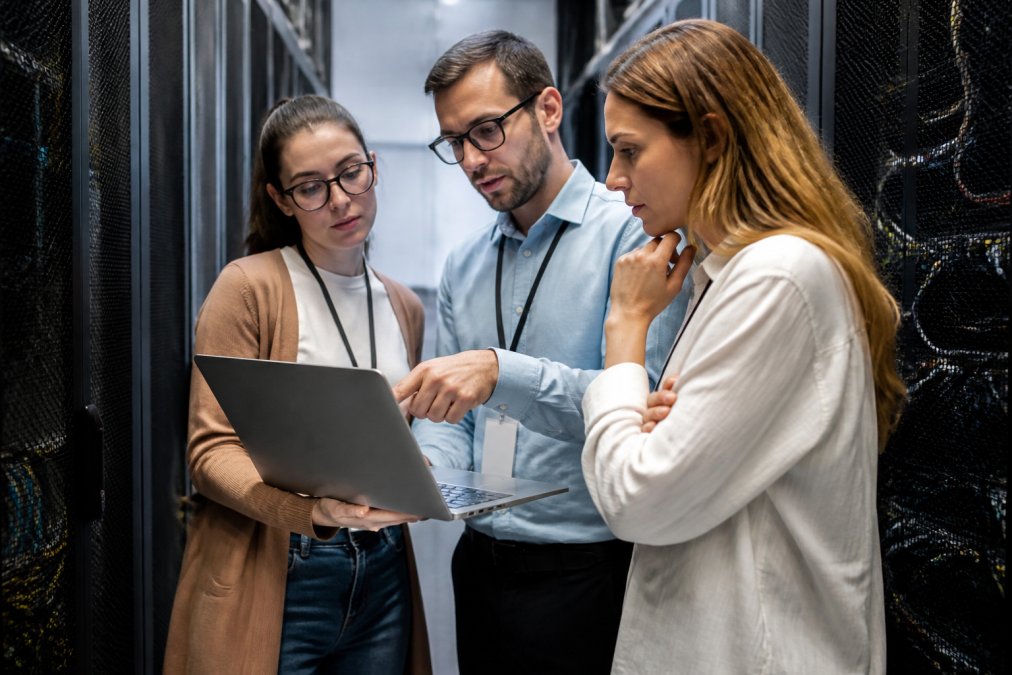 IT team members in a modern data center reviewing system architecture diagrams displayed on multiple monitors, pointing at legacy and modern infrastructure components while discussing integration strategy