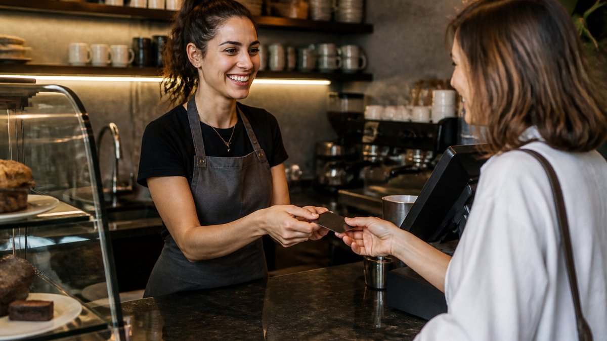 Barista handing a digital loyalty membership card or tablet to a customer at a cafe counter during checkout, with warm cafe lighting and a register in background
