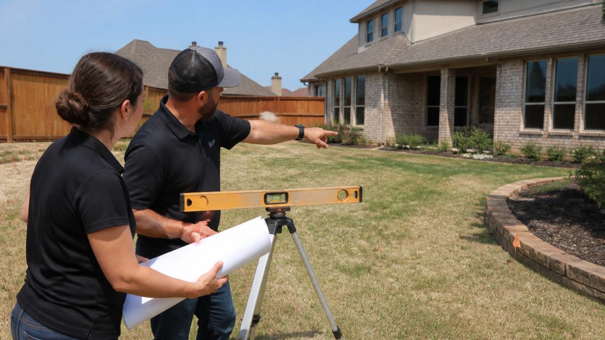 A landscape designer and a pool builder standing in a residential backyard holding rolled site plans, pointing toward the house and existing fence line while measuring the slope with a level
