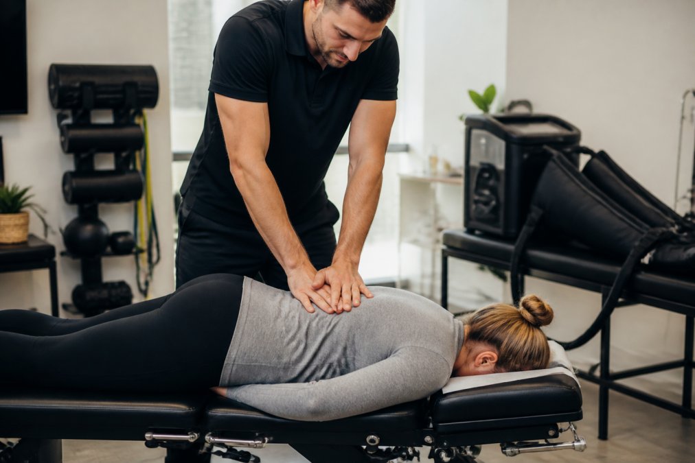 Chiropractor performing spinal adjustment on patient in modern clinical treatment room with professional lighting, recovery equipment visible in background