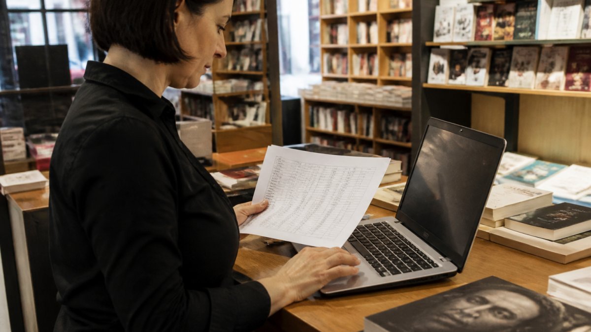 A bookseller at a desk in an independent bookstore, comparing a printed customer list with a laptop screen showing a database import, shelves of books softly blurred in the background