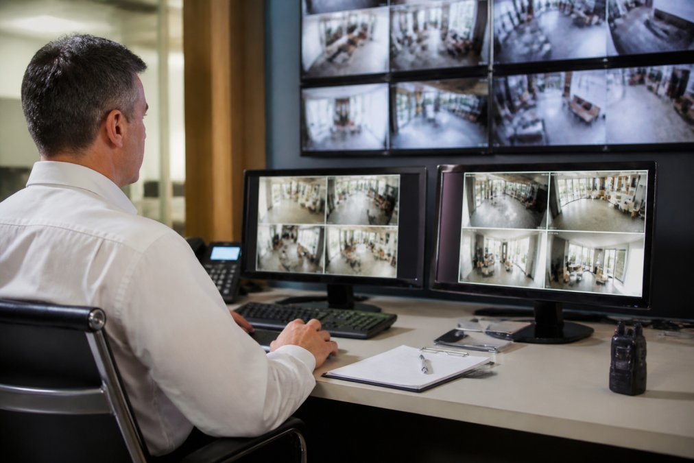Facility manager reviewing security system monitoring dashboard on a desktop computer in a control room, with multiple camera feeds visible on screens in professional lighting