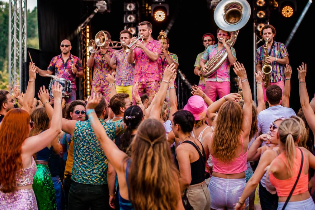 Een energieke brassband die optreedt op een podium terwijl het publiek uitbundig danst en meebeweegt, met kleurrijke kleding en instrumenten in close-up