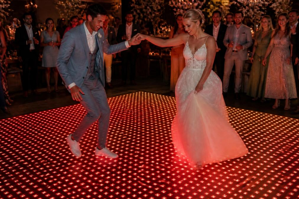 Bruidspaar dansend op een verlichte dansvloer met warm rood-oranje LED-spectrum, gasten kijkend vanaf de zijkanten, feestelijke sfeer met directe reactie van lichten op hun voetdrukken