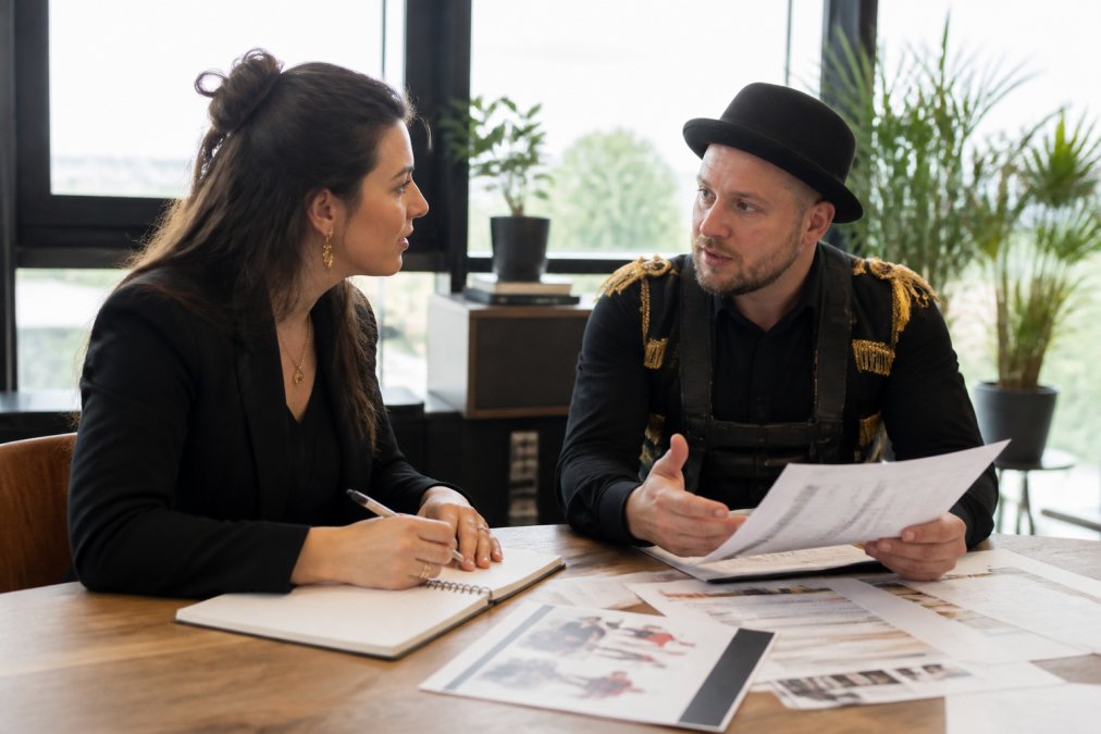 Event planner in conversation with a performer, both reviewing notes and technical specifications on a table with planning documents visible, professional office setting with natural lighting
