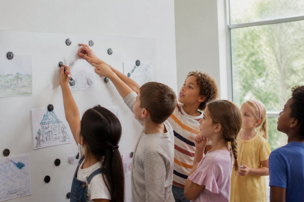 Diverse group of primary school students collaborating around a magnetic whiteboard wall, pointing and writing together with engaged expressions, natural classroom lighting streaming through windows