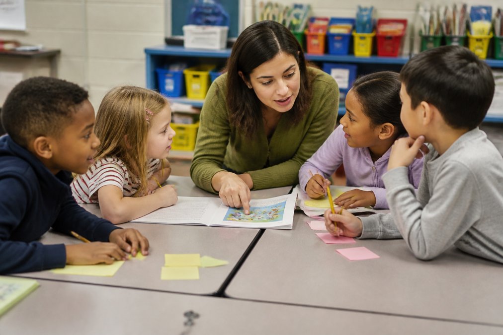 An elementary teacher kneeling beside a small group of students at a classroom table, pointing at a printed story booklet while the children write on sticky notes and talk with each other
