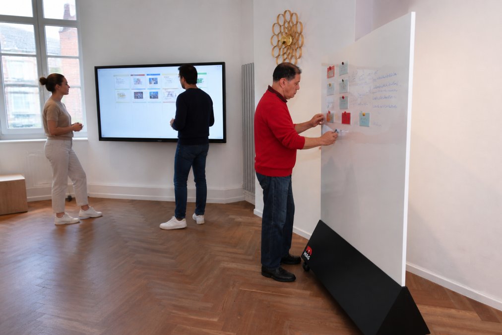 Professional office space showing a magnetic interactive wall on one side with colour-coded project cards and handwritten notes, and a large digital display screen on the other side with team members using both surfaces, bright natural lighting from windows