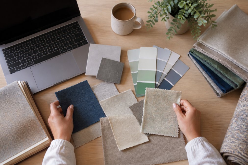 Overhead shot of hands arranging fabric swatches and paint chips on a light oak table beside a laptop showing a style quiz