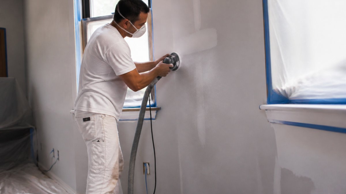 Professional painter sanding drywall, a key step affecting interior painting cost, with drop cloths and masking tape.