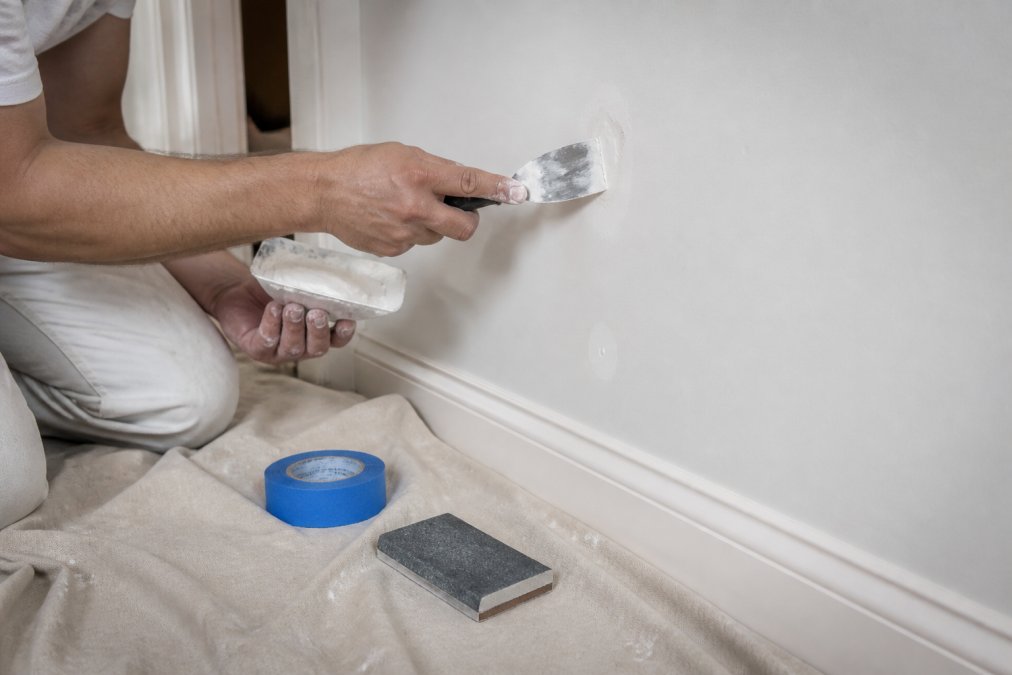 A close-up of hands using a putty knife to apply spackle to a small hole in a white interior wall, with a sanding block and painter's tape visible on a drop cloth below