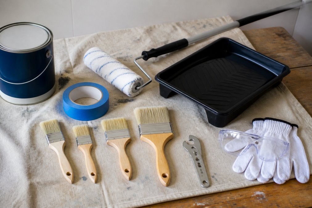 Organized collection of interior painting supplies arranged on a canvas drop cloth: synthetic bristle brushes in various sizes, 9-inch paint roller with extension pole, blue painter's tape, paint tray, paint can opener, and protective gear (gloves, safety glasses) on a wooden workbench