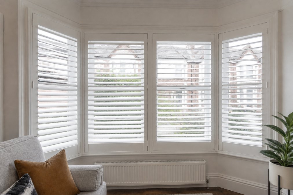 Close-up of white plantation shutters fitted across a Victorian-style bay window in a bright, well-decorated living room, showing the angled panels meeting neatly at each bay section with clean mitred joins at the frame corners
