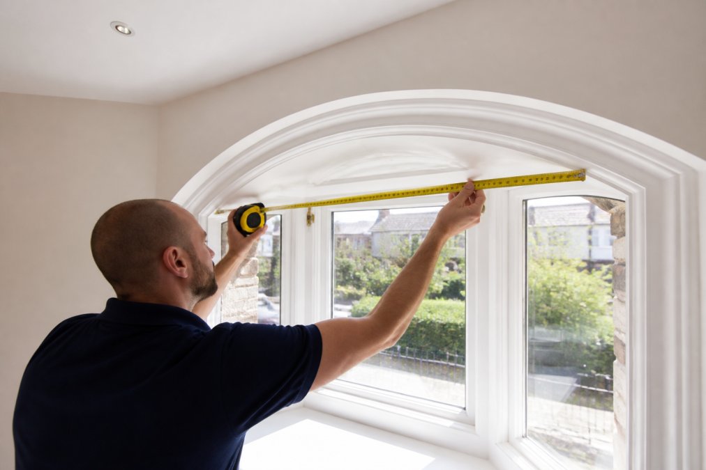 Close-up of homeowner's hand measuring bay window frame with tape measure, showing complexity of arched window geometry with natural daylight revealing frame details