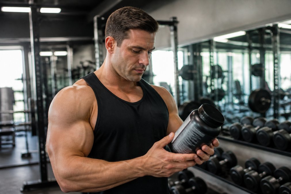 A muscular athlete in a gym holding a supplement bottle, examining the label with focused expression, dumbbells and training equipment visible in the background