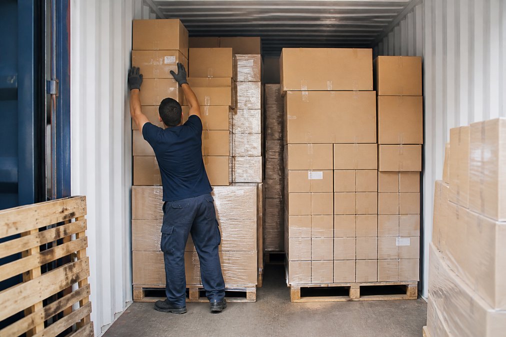 Warehouse worker organizing palletized cargo in a shipping container, stacking boxes strategically to maximize space use, with natural overhead lighting