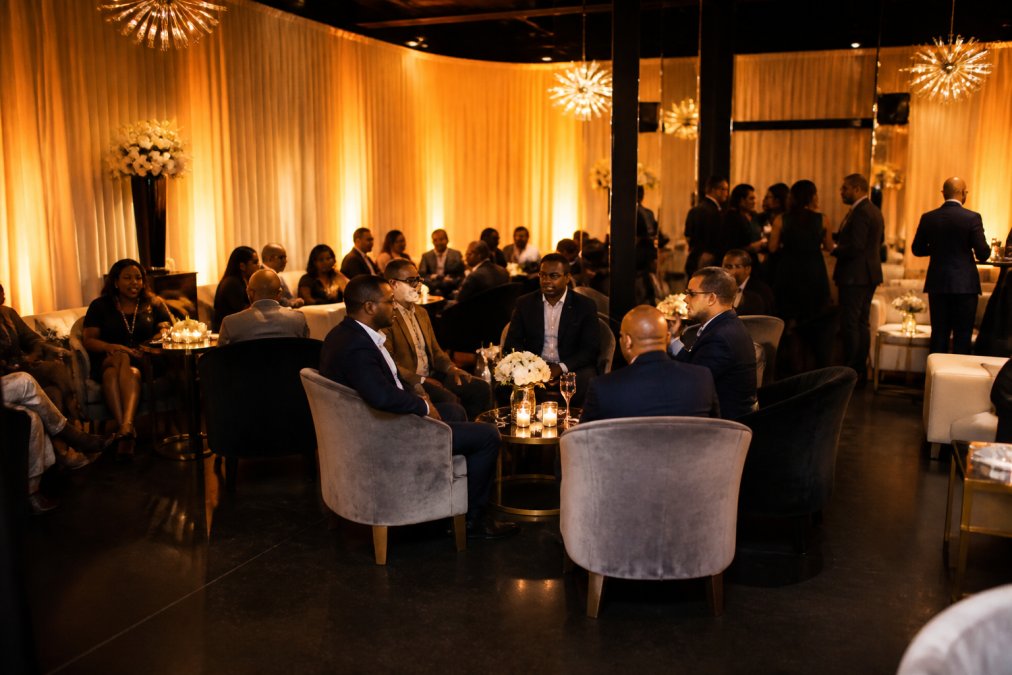 Elegantly decorated intimate event lounge with soft amber up-lighting, plush velvet lounge chairs, and draped ivory walls set for a corporate gathering of around 30 guests seated around low cocktail tables