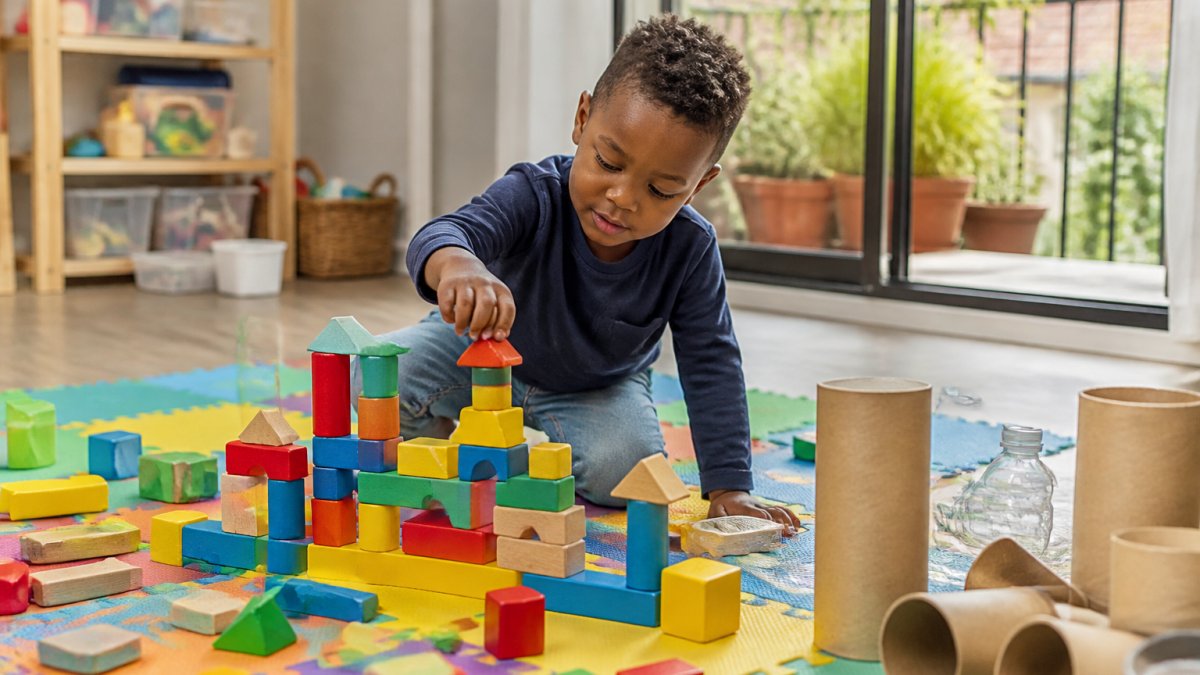 Young child concentrating while stacking coloured wooden blocks and construction pieces on a play mat, with cardboard tubes and household materials visible nearby in natural daylight