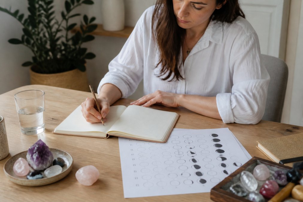 Frau schreibt konzentriert in ein Journal, neben ihr liegen ein Mondkalender und handverlesene Kristalle auf einem hellen Holztisch