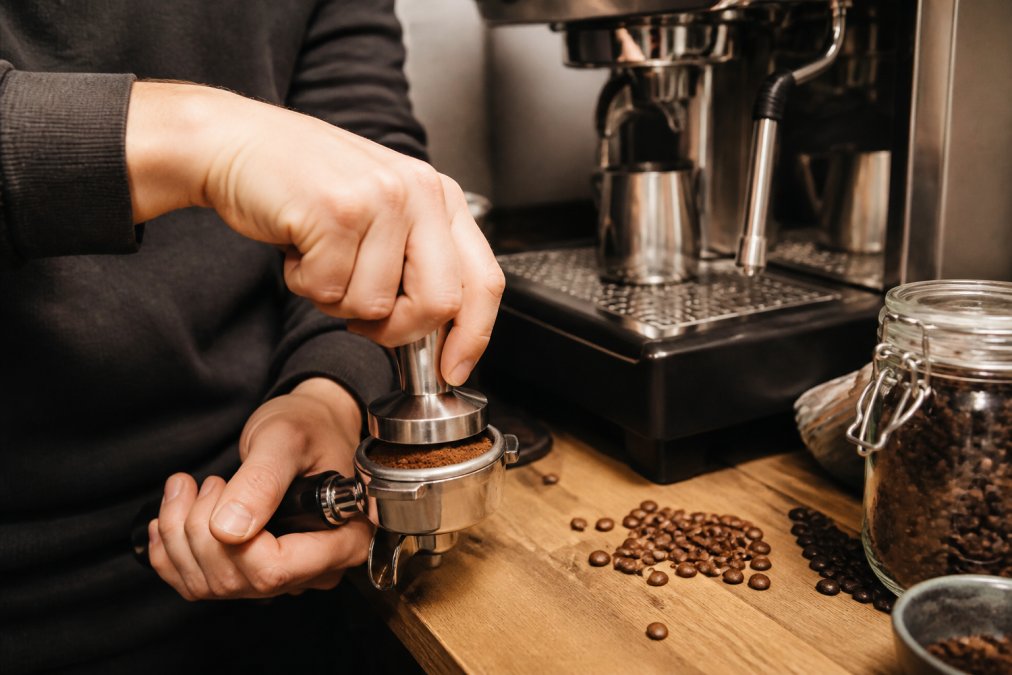 Barista's hands tamping espresso grounds in portafilter with espressomaskine visible in background, coffee beans scattered on counter, warm kitchen lighting