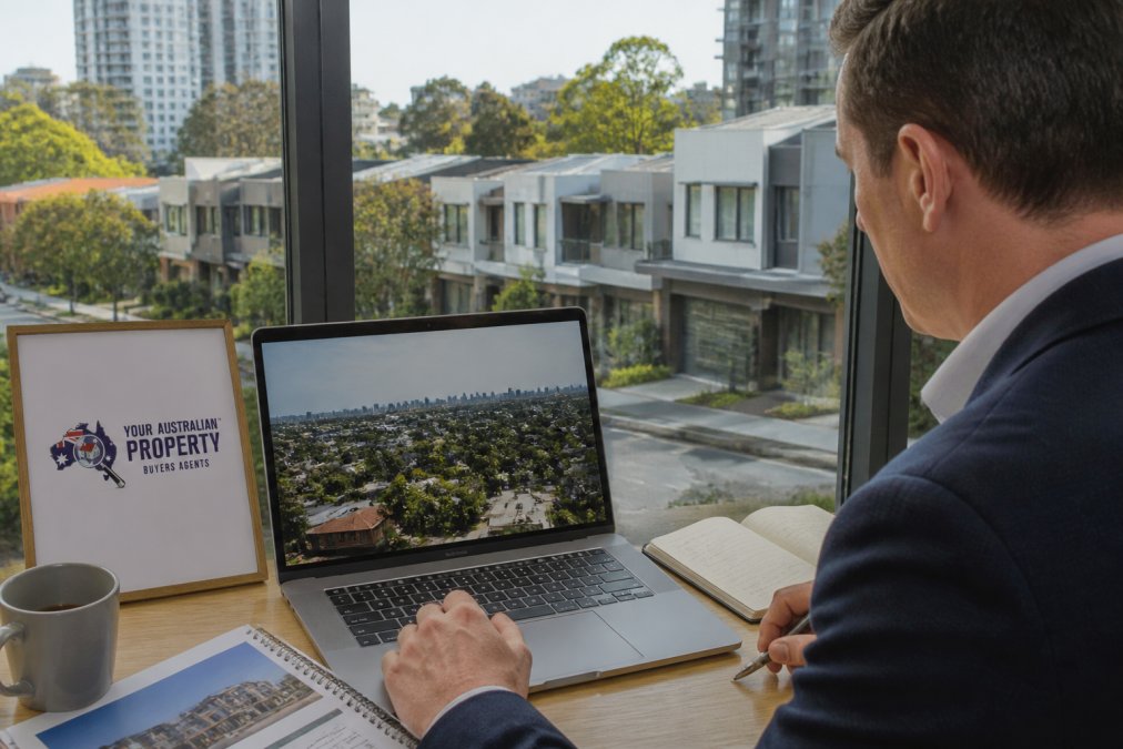 Property investor reviewing suburb data and growth forecasts on laptop at desk with Melbourne neighbourhood street view through window showing modern townhouses and apartment buildings