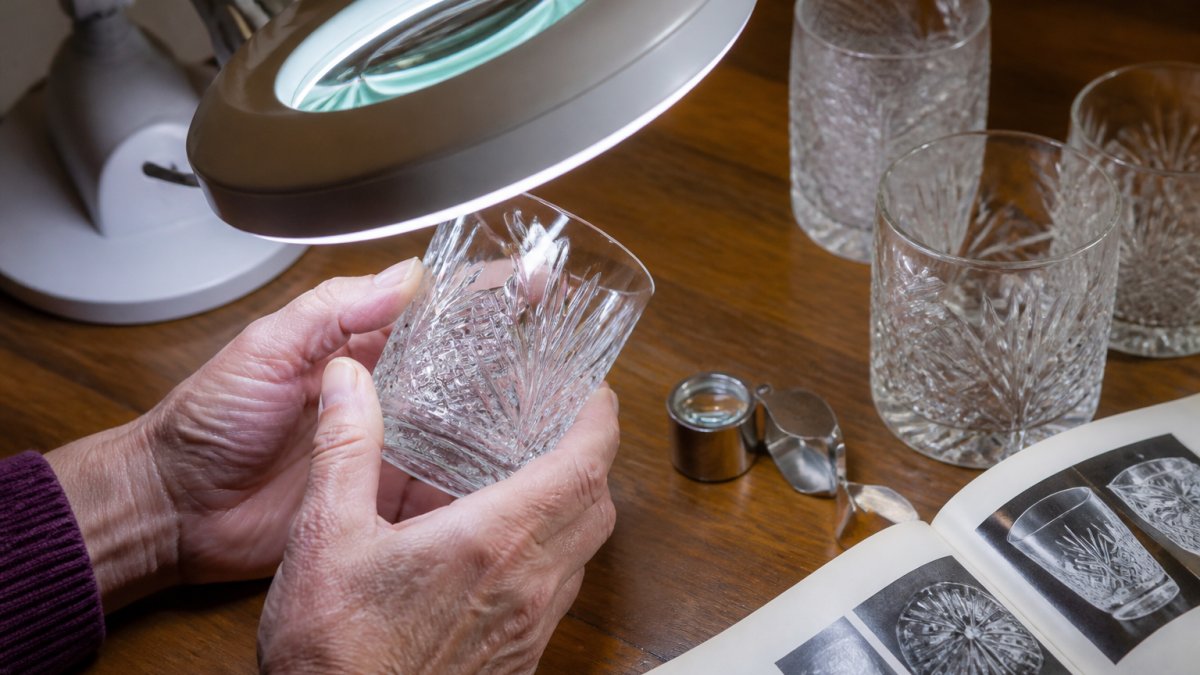 Close-up of hands holding a vintage cut-glass tumbler under a magnifying lamp on a wooden table, with a jeweler's loupe and a reference book nearby