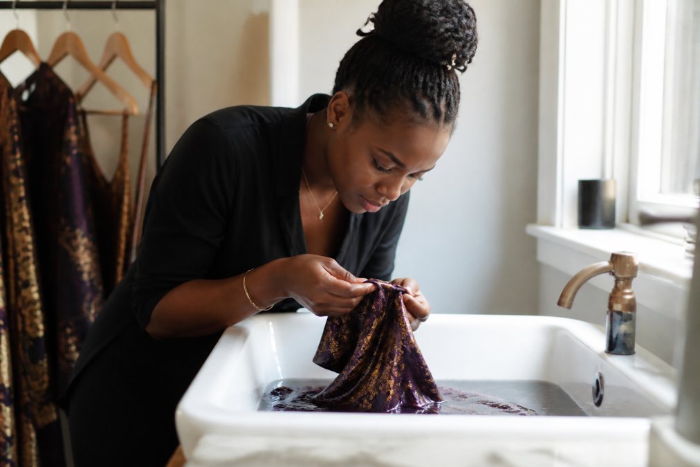 Woman carefully hand-washing a luxurious jewel-toned occasion dress in a pristine white sink, inspecting delicate seams with focused attention, natural daylight streaming through a window