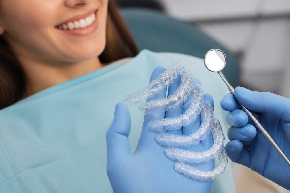 Close-up of transparent clear aligner trays arranged in progression showing the incremental tooth movement stages, with a dental mirror and patient's teeth visible in soft clinical lighting