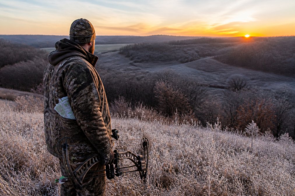 A hunter in layered camo glassing across a rolling Iowa timber edge at first light, bow in hand, frost on the grass and a folding map tucked in a jacket pocket