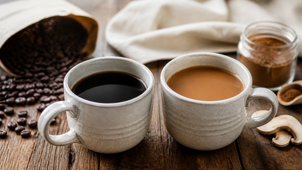 Close-up of two mugs side by side on a wooden counter, one filled with dark black coffee and one with a lighter tan mushroom coffee blend, with a bag of whole coffee beans and a small jar of mushroom powder visible in the background