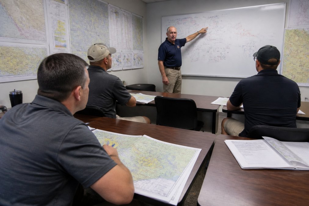 Students seated in a small classroom reviewing aviation charts and weather maps at desks, with an instructor pointing to a whiteboard covered in flight operations notes, under bright fluorescent lighting