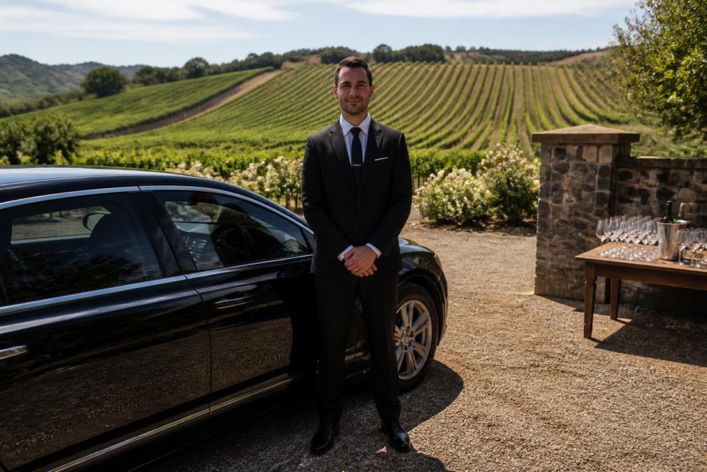 Professional chauffeur in formal attire standing beside a luxury sedan at a vineyard entrance, with wine-tasting glasses visible on a wooden table in the background and rolling vineyard rows stretching across the landscape