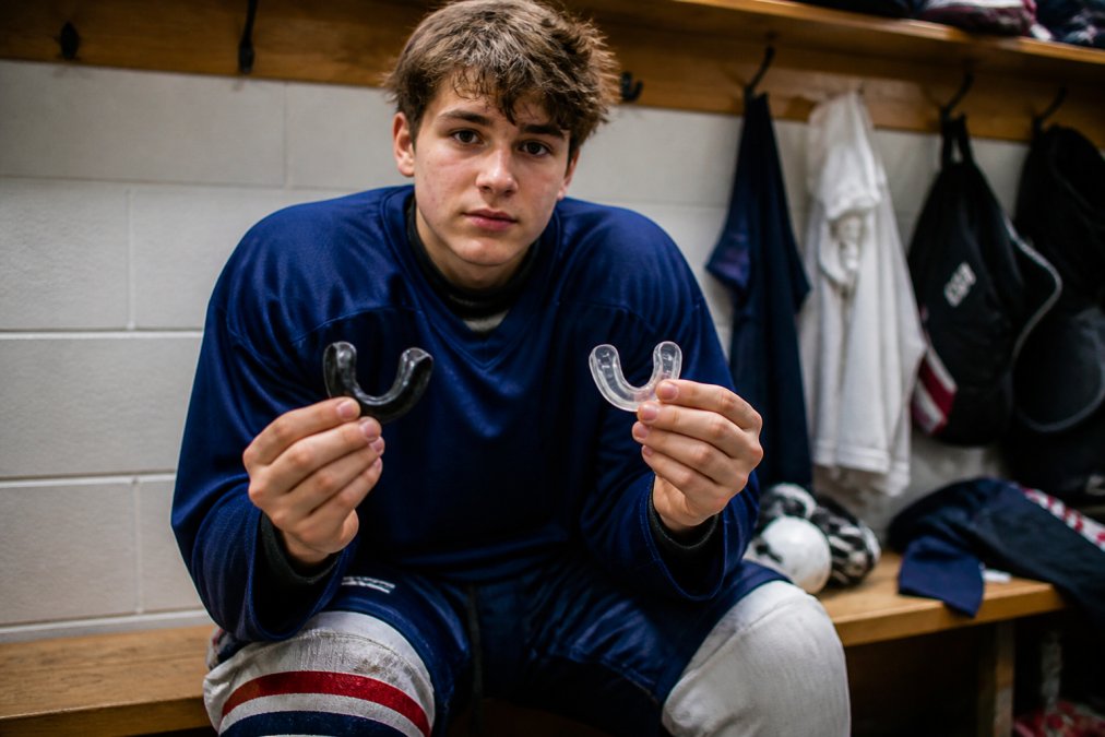 Teenage hockey player comparing a black custom mouthguard to a clear boil-and-bite guard on a locker room bench