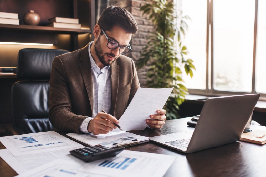 Real estate investor in business casual attire reviewing property financial statements and loan documents at a modern office desk, with a calculator, laptop, and market analysis papers visible under warm office lighting