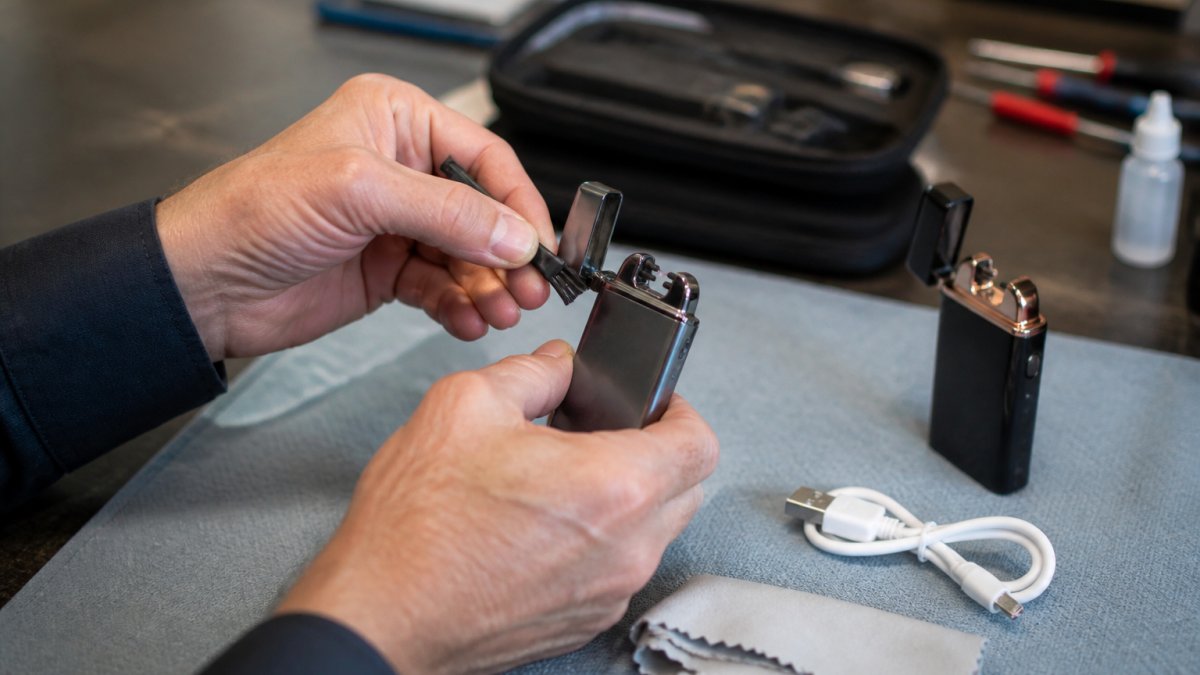 Hands using a small brush and cloth to clean the arc prongs of an open electric plasma lighter on a workbench