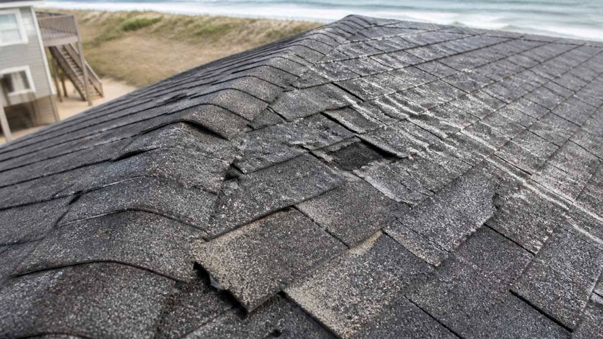 Close-up of roof shingles showing visible curling, missing pieces, granule loss, and salt-spray damage on coastal home