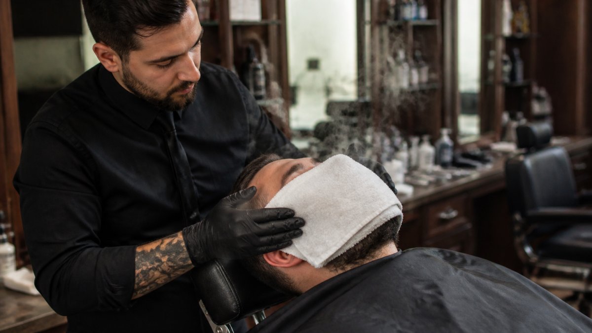 Professional barber applying steaming hot towel to client's face during traditional shave, with visible steam and client in relaxed reclined position in modern barbershop setting