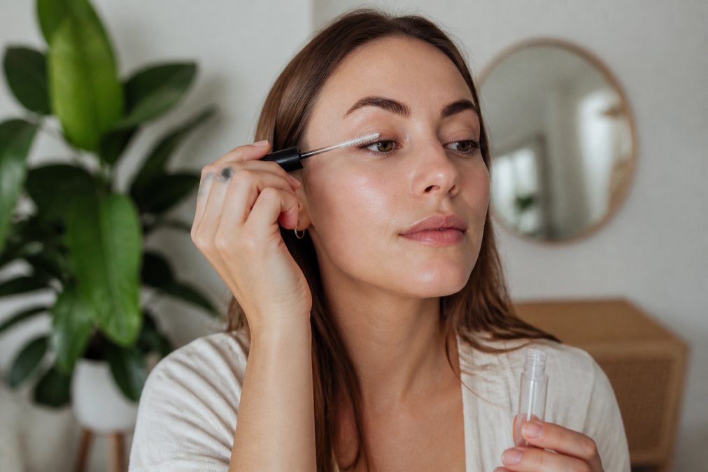 Woman applying a conditioning serum or nourishing aftercare product to her lashes with a small applicator brush, showing proper maintenance technique in natural soft lighting
