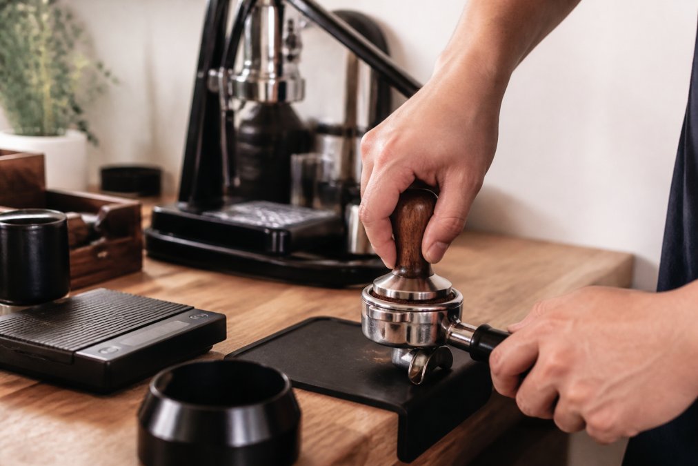 Close-up of hands tamping freshly ground coffee into a portafilter on a wooden countertop, with a manual lever espresso machine visible in the background and a small scale nearby