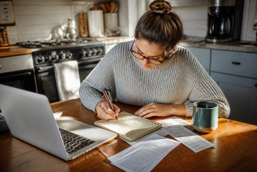 A person sitting at a kitchen table with a laptop open to a bank statement, a notepad with a handwritten list of subscriptions, and a coffee cup nearby, in warm morning light suggesting a focused personal finance review session