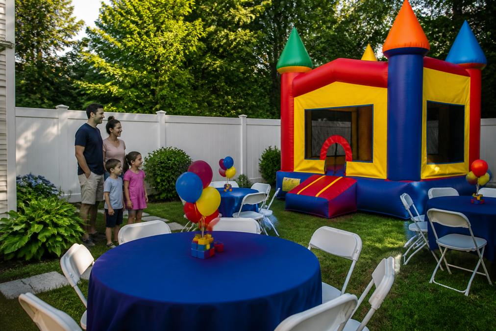 Professional event setup showing a bounce house installed in a backyard with coordinated tables, chairs, and linens arranged for a children's party, with a family watching and smiling in the background, afternoon sunlight