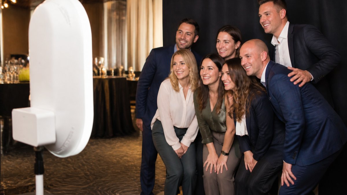 Professionally dressed colleagues posing and laughing inside a sleek open-air photo booth at a corporate reception, branded backdrop visible, warm event lighting