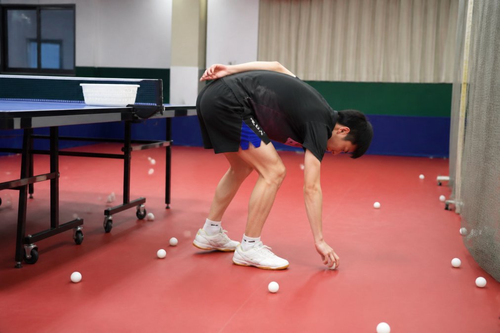 Table tennis player bending over repeatedly to pick up scattered balls on the floor during training session, showing poor posture and repetitive strain on lower back in brightly lit training facility