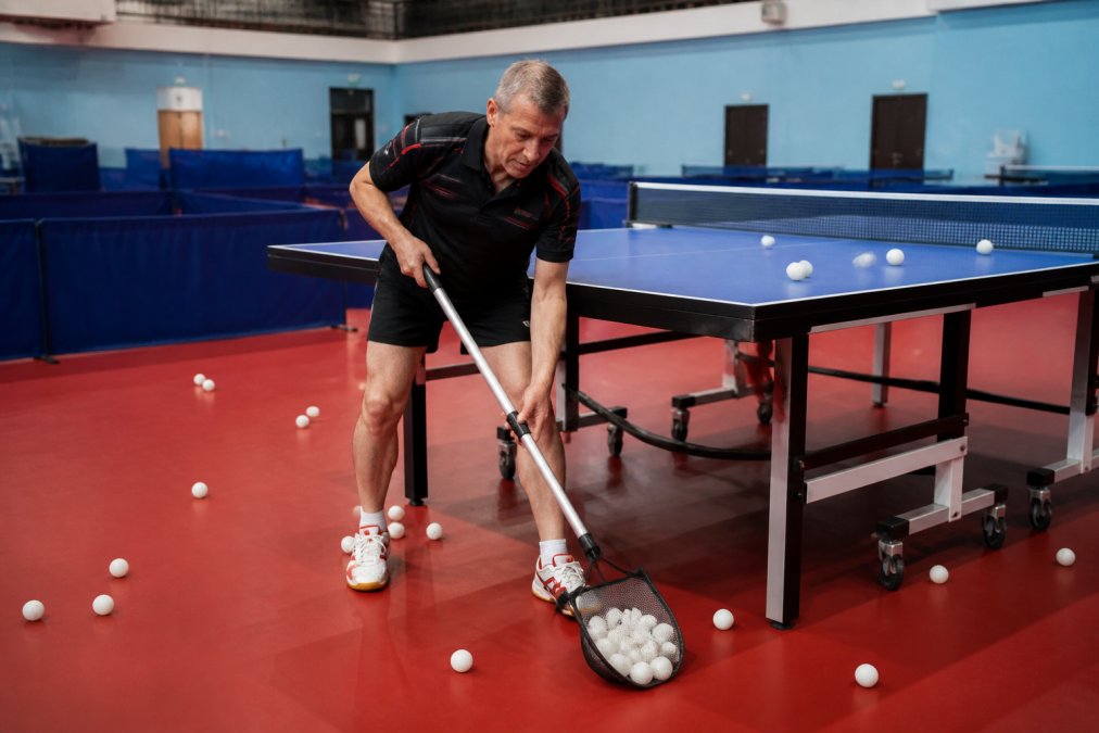 Experienced coach using ball collector to efficiently gather multiple balls during multi-ball training session at professional table tennis table, with balls scattered across floor and table surface, natural gym lighting