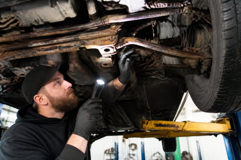 Professional mechanic examining the undercarriage of a used vehicle with a flashlight and inspection tools, showing rust and corrosion details on suspension components