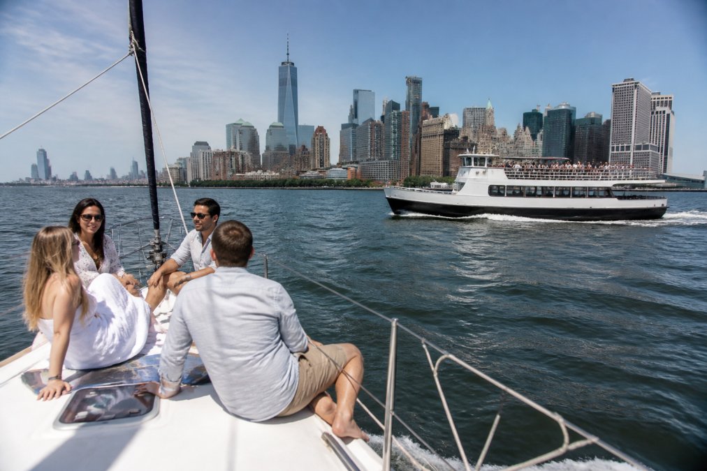 A small group of friends relaxing on the deck of a private sailboat with a city skyline in the background, while a larger crowded tour boat passes in the distance