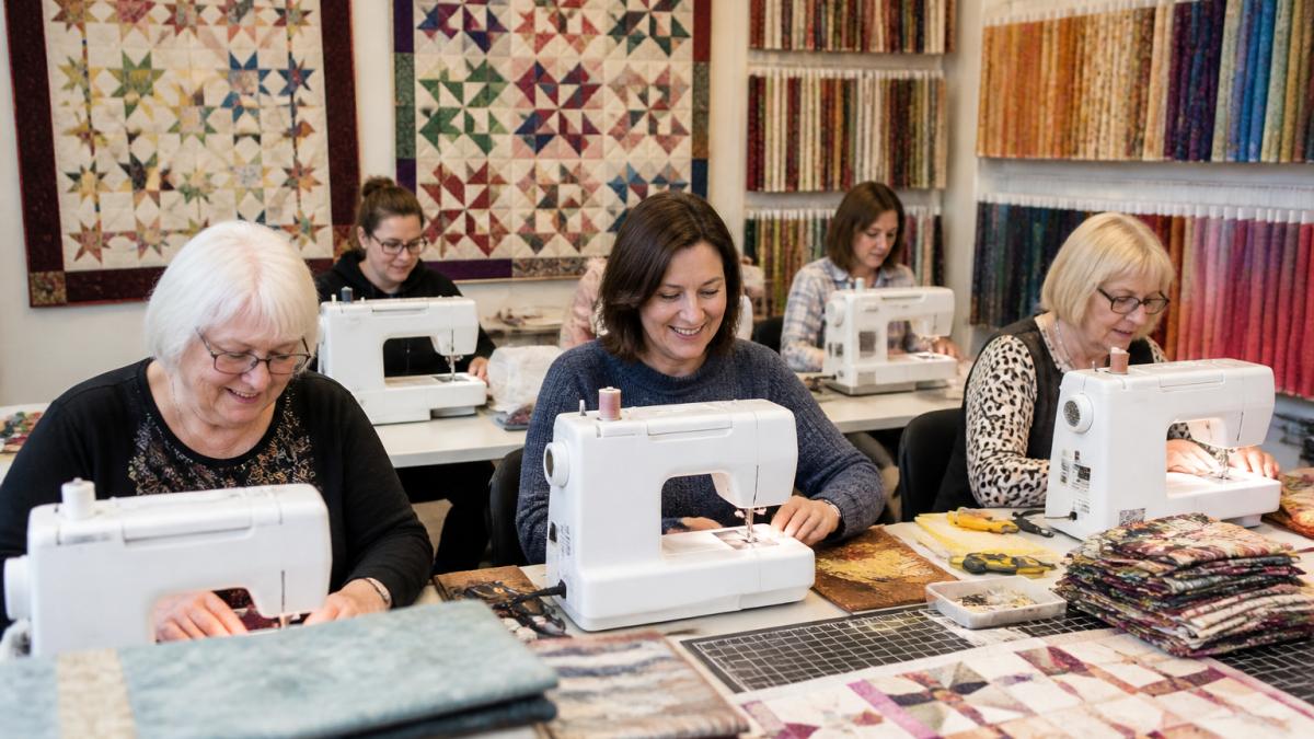 A small group of quilters of varying ages seated at sewing machines in a bright, welcoming quilt shop classroom, with fabric swatches and a finished quilt sample on the wall behind them