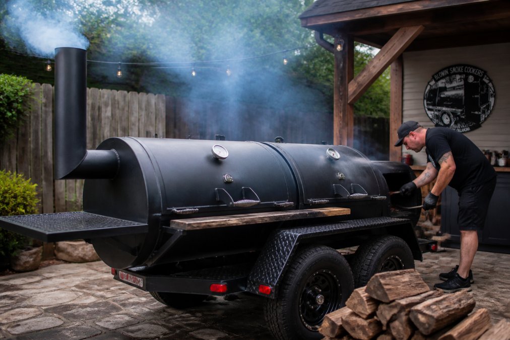 Heavy-duty offset professional smoker with thick steel on a patio as a person tends the firebox.