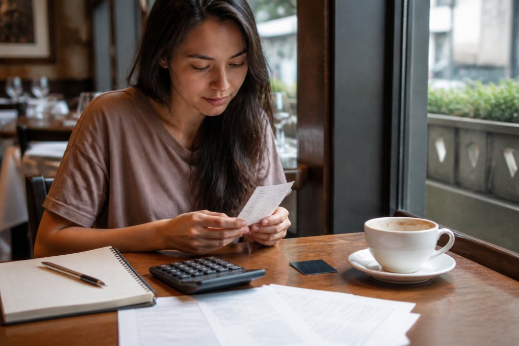 A person sitting at a restaurant table reviewing a paper receipt next to a loyalty card, with a coffee cup and soft natural light from a nearby window